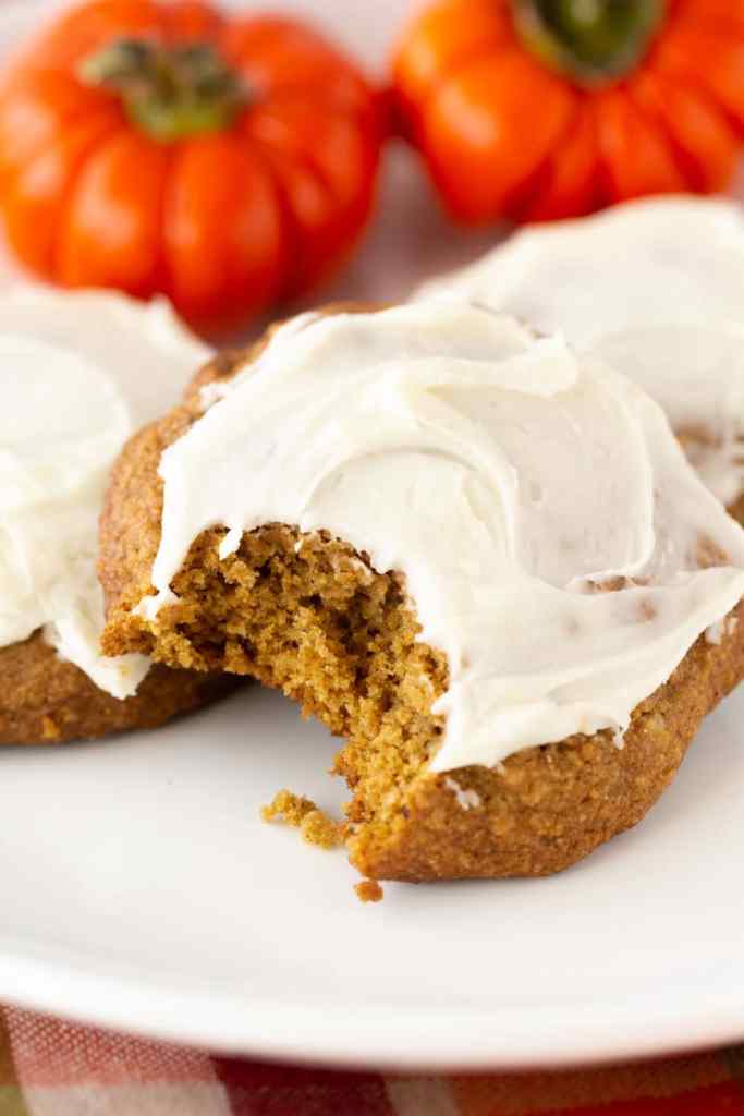 A close-up of a pumpkin cookie with cream cheese frosting, showing a bite taken out, with small orange pumpkins in the background.