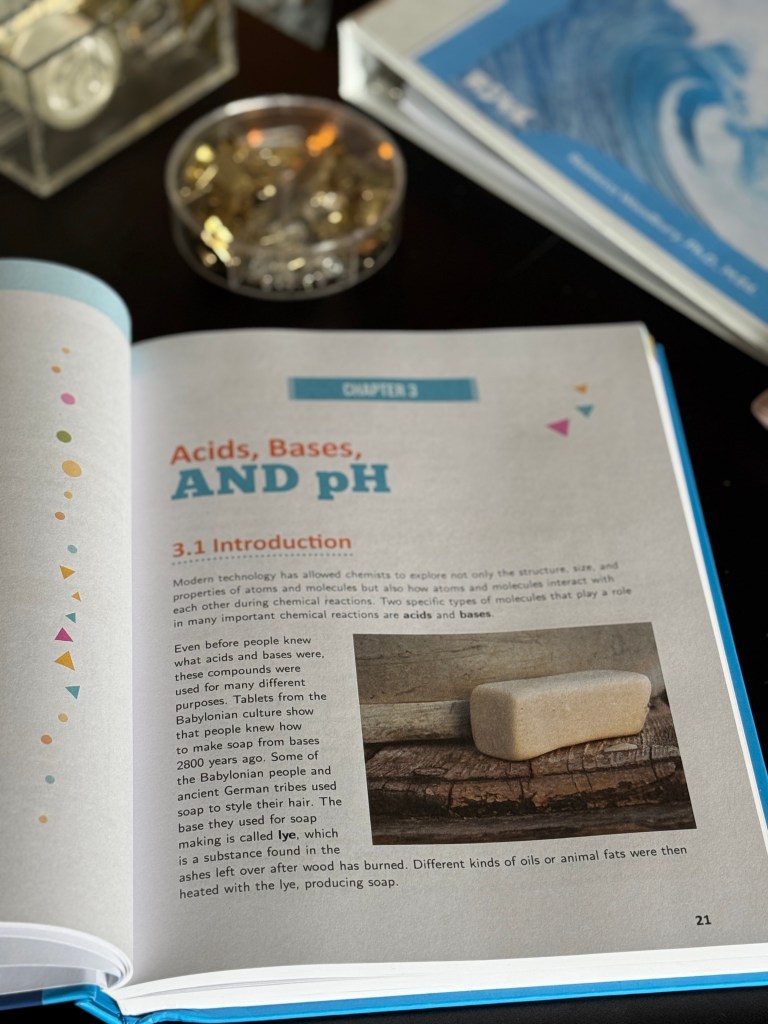An open textbook page titled 'Acids, Bases, AND pH' featuring colorful graphics and a stone example of soap on a wooden surface.
