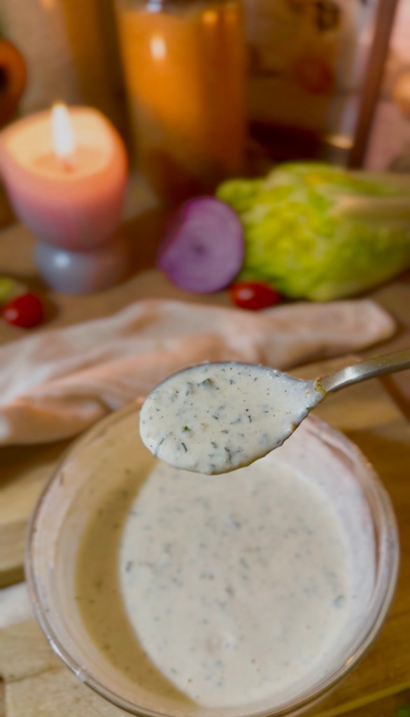 A spoonful of creamy homemade ranch sauce being lifted from a glass bowl, with fresh vegetables and a lit candle in the background.
