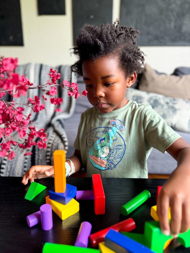 A young child focuses on building with colorful geometric blocks on a table, surrounded by flowers and soft furnishings.