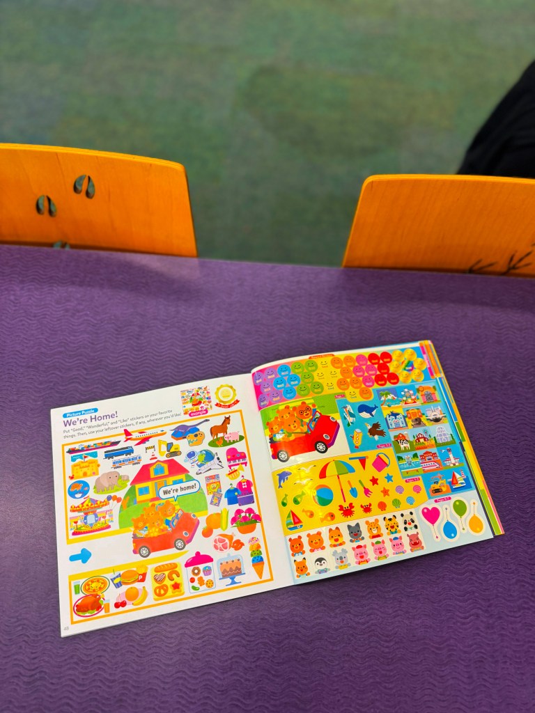 A children's activity book opened to a page featuring colorful stickers and illustrations, including a red car and various playful images. The background shows a purple table and wooden chairs.