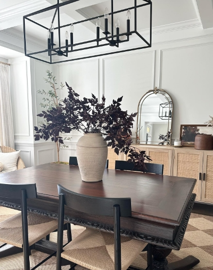 Dining room featuring a dark wooden table with a large vase of dark foliage as a centerpiece, surrounded by black chairs and a warm light fixture above.