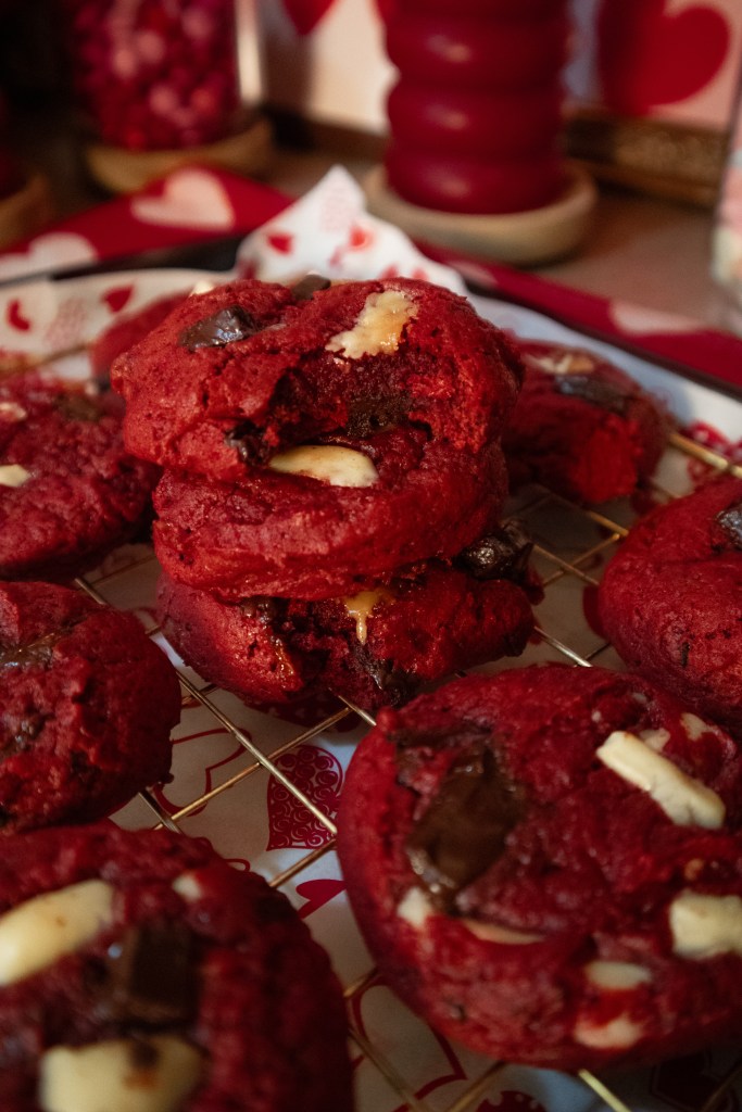 A close-up of a stack of red velvet cookies, some with chocolate chunks and white chocolate pieces, on a decorative tray with heart patterns.