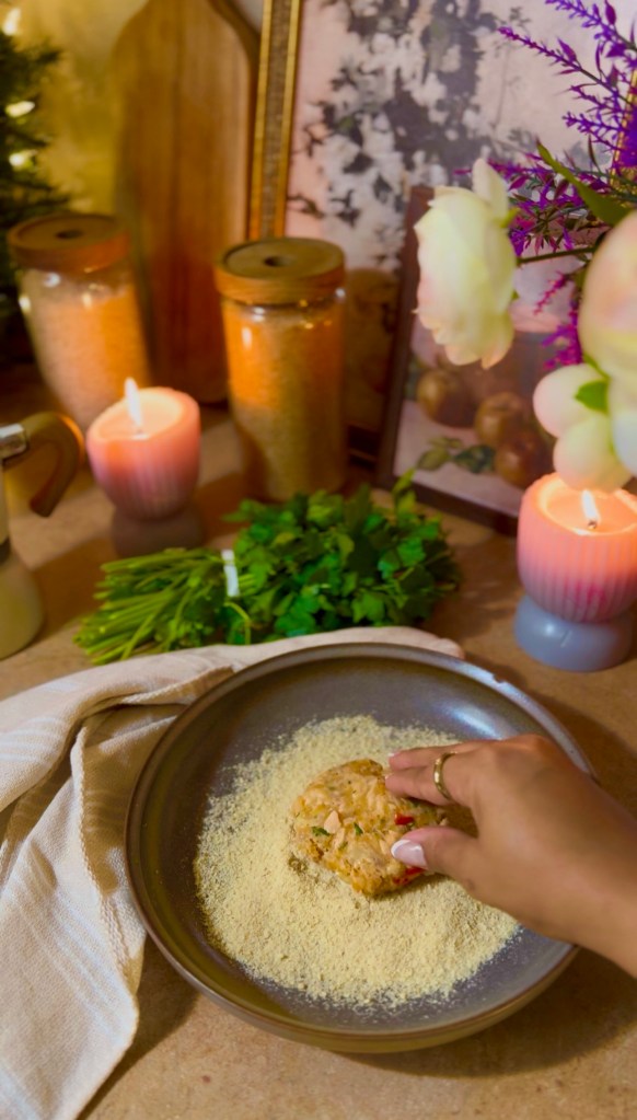 A hand pressing a salmon patty into breadcrumbs on a grey plate, surrounded by jars of spices, candles, fresh herbs, and a decorative background.