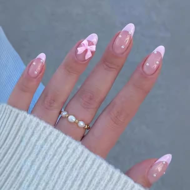 A close-up of a hand with long, pink French tip nails, featuring a pink bow accent on one nail. The hand is adorned with a delicate pearl and gold ring, and is partially covered by a light-colored sweater.