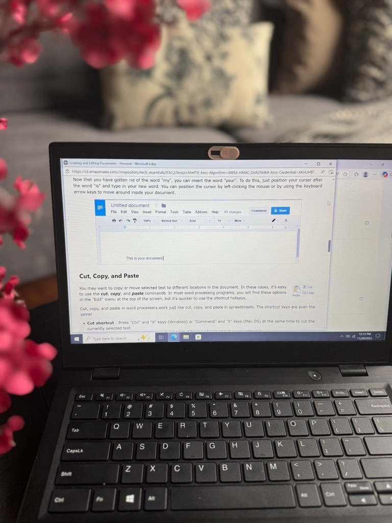 A close-up view of a laptop displaying a word processing document titled 'Creating and Editing Documents - Personal - Microsoft Edge.' The screen shows instructions on how to cut, copy, and paste text in a word processor, with additional text and a cursor visible. In the foreground, there are pink flowers blurred out.