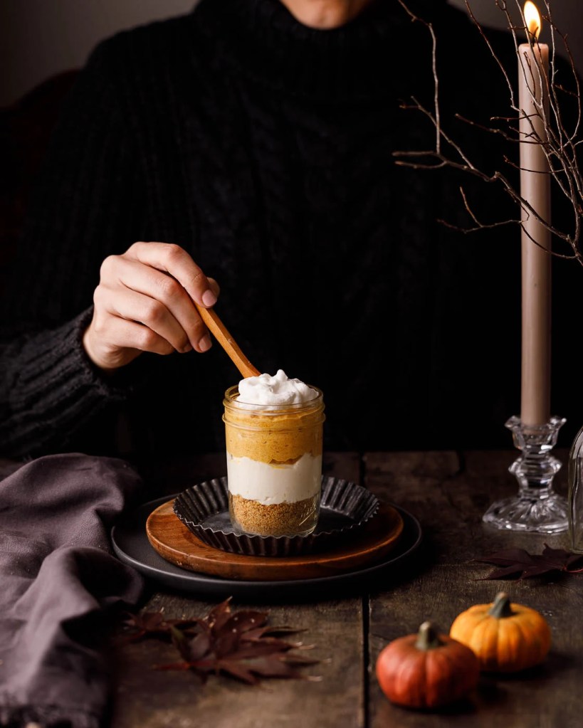 A person holding a spoon above a layered dessert in a jar, featuring pumpkin puree and whipped cream, on a rustic wooden table decorated with small pumpkins and a candle.