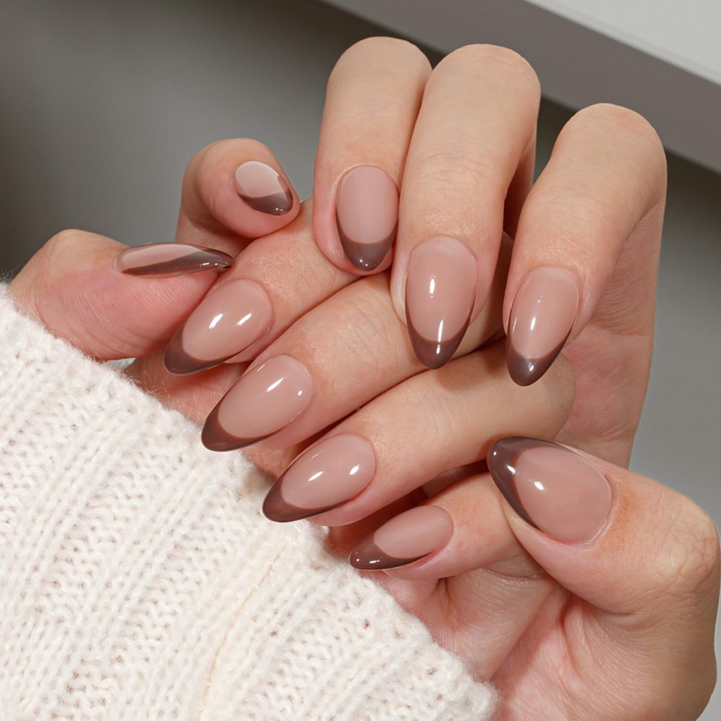 Close-up of manicured hands with almond-shaped nails featuring a nude base and dark brown tips, holding each other. One hand is partially covered by a cozy knitted sweater.