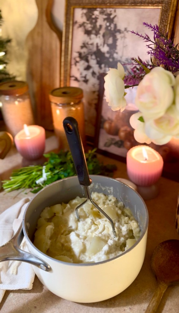 A pot of freshly mashed potatoes with a potato masher resting on the edge, surrounded by decorative elements including candles, flowers, and jars.
