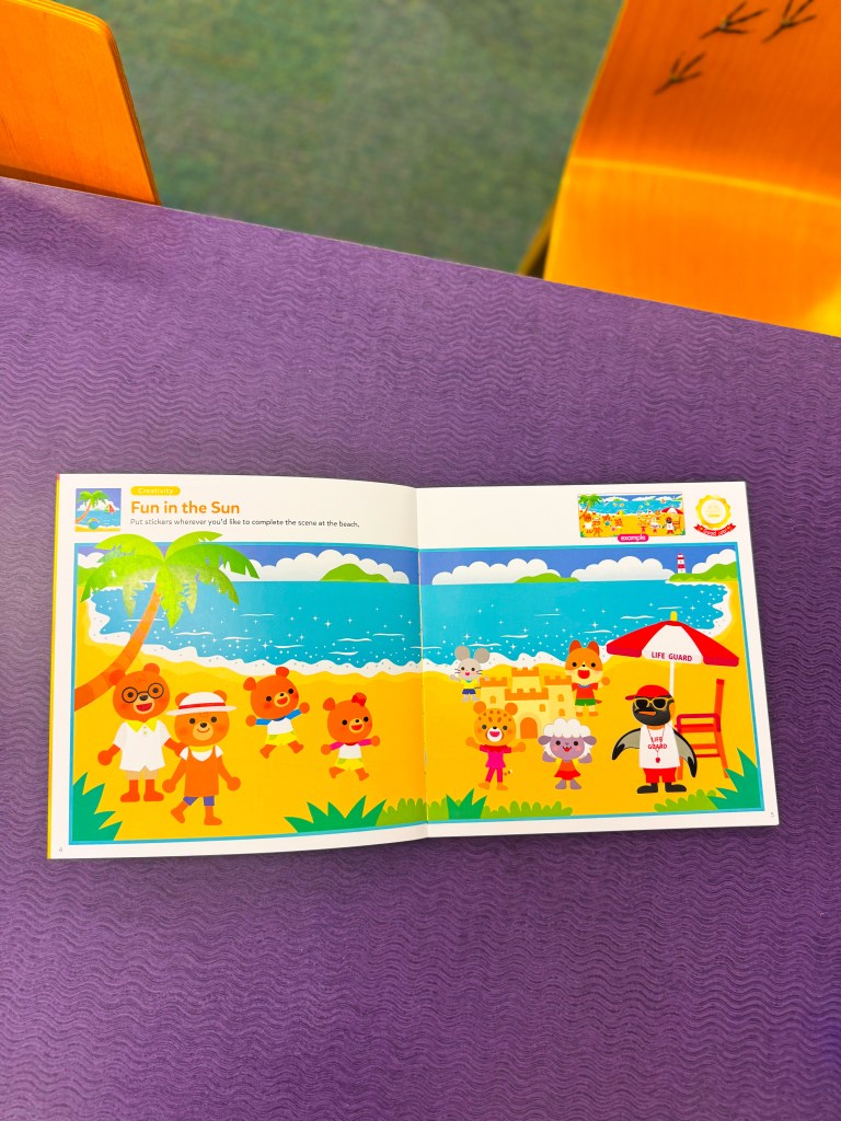 A children's activity book open to a colorful beach scene titled 'Fun in the Sun', featuring cartoon animals enjoying the beach, with various playful elements like palm trees, surf, and a lifeguard.