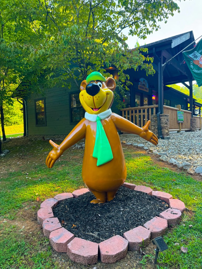 A colorful statue of a bear wearing a green hat and scarf, standing in a grassy area surrounded by a circular stone border, with a rustic building in the background.