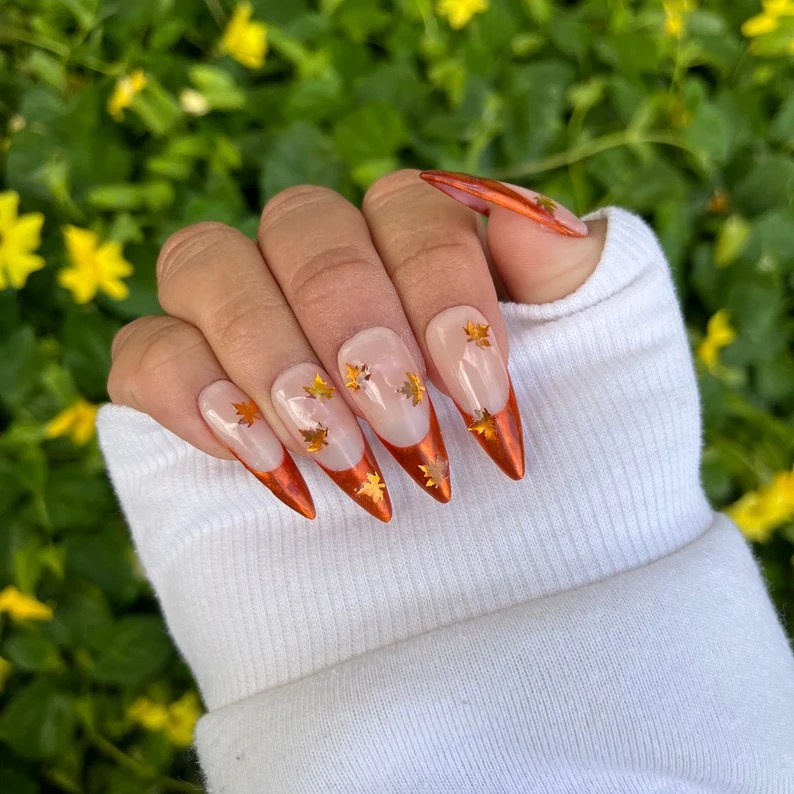 A hand with long, pointed nails featuring a burnt orange tip and gold leaf accents against a clear background, resting on a white sweater in front of green foliage.