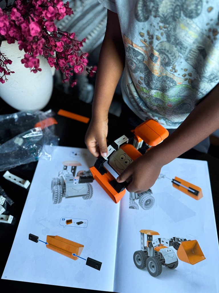 A child assembling a toy robot vehicle using building blocks, with an instruction manual open beside them, surrounded by pink flowers in a vase.