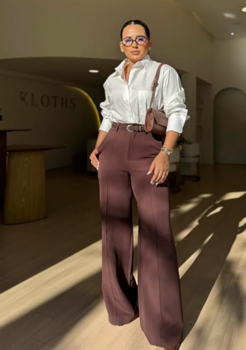 A woman wearing a white shirt and brown wide-leg trousers stands in a well-lit space, accessorized with glasses and a small bag.