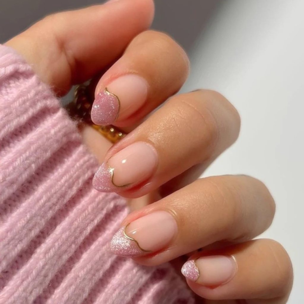 A close-up of manicured nails featuring a nude base with pink glitter tips and gold accents, positioned against a soft pink knitted sweater.