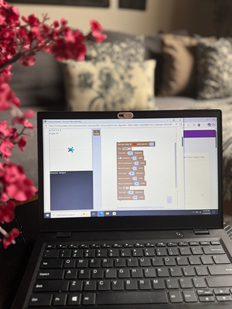 A laptop screen displaying a coding interface with blocks for programming commands, showcasing an educational tech lesson, alongside a blurred background of floral decor.