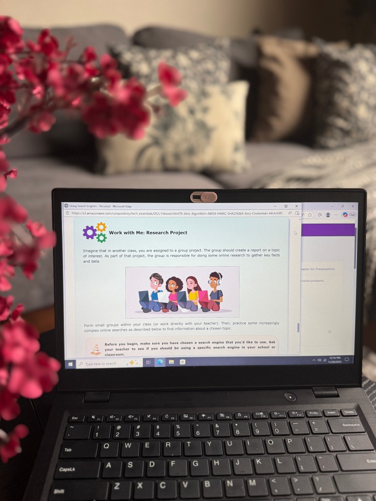 A laptop displaying an online lesson titled 'Work with Me: Research Project' with an illustration of four diverse children engaged in discussion. In the background, a cozy living room setting is visible, featuring decorative pillows and a flowering plant.