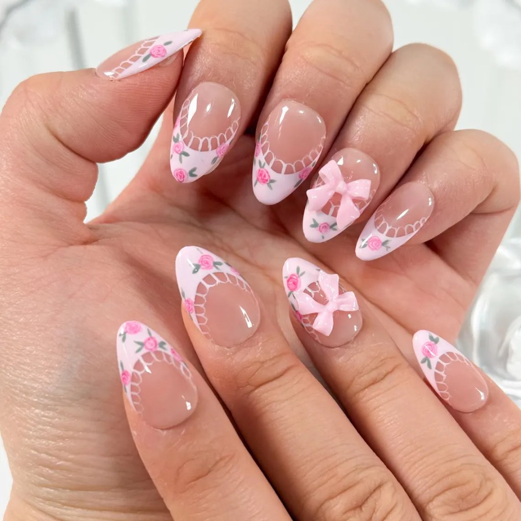 Close-up of elegantly manicured nails featuring a pink and white design with floral patterns and pink bows, displayed on a hand.