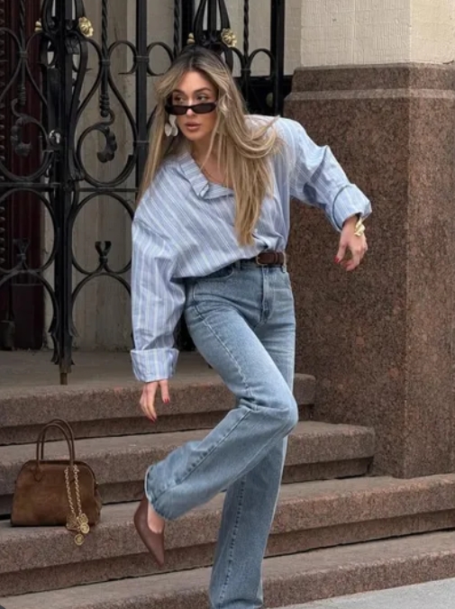 A stylish woman in a striped blue oversized shirt and high-waisted jeans poses outside, complemented by sunglasses and a chic handbag.