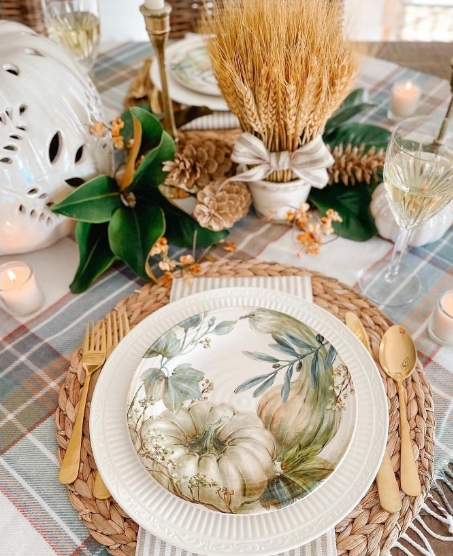 A beautifully arranged fall dining table featuring a decorative plate with pumpkin artwork, golden cutlery, and a centerpiece of wheat and floral elements. The table is set against a plaid tablecloth with soft candlelight enhancing the cozy atmosphere.