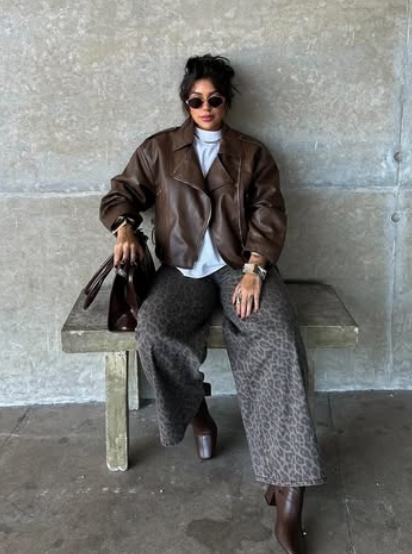 A woman seated on a bench wearing a brown leather jacket over a light gray top, paired with wide-leg leopard print pants and sunglasses. She accessorizes with a stylish handbag and jewelry against a concrete background.