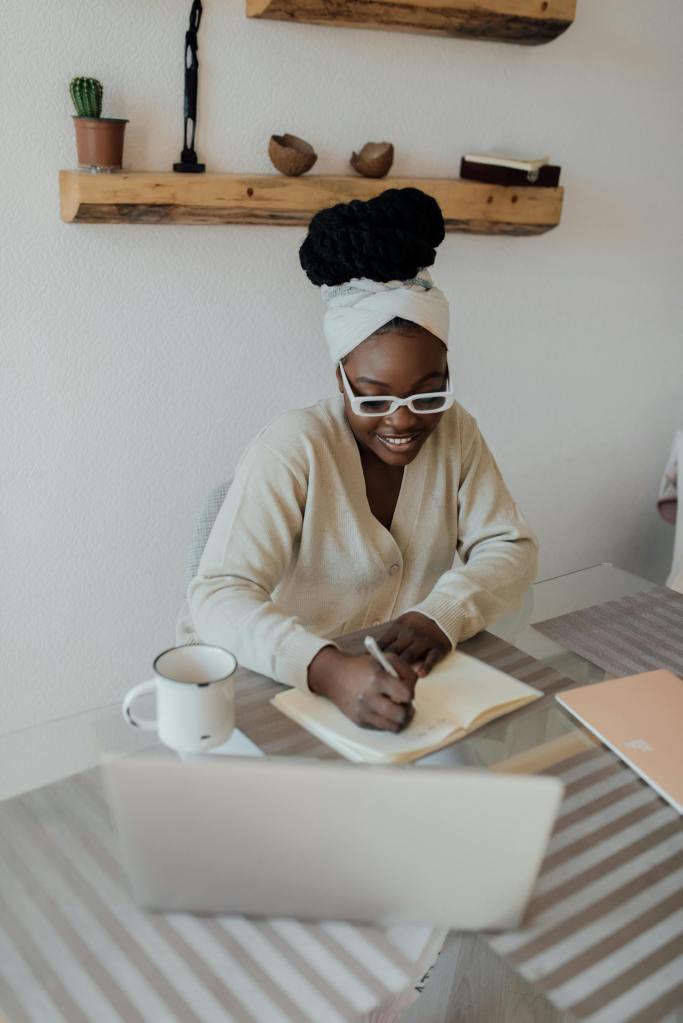 A person with a headwrap and glasses smiles while writing in a notebook at a table, with a laptop and mug nearby.