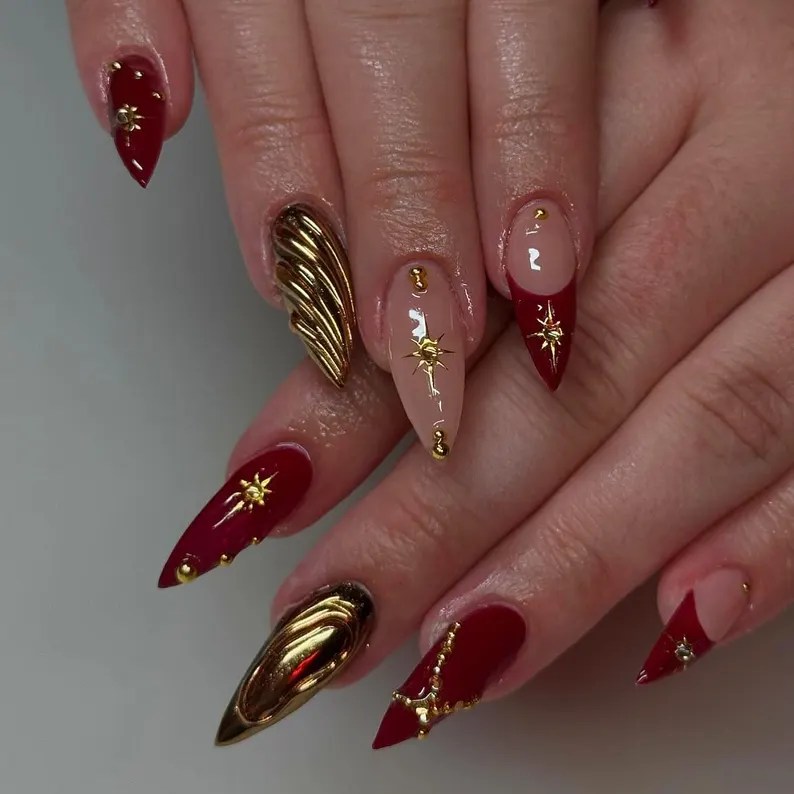 A close-up view of hands with long, pointed nails in red and gold designs, featuring intricate detailing like stars and a wing motif.