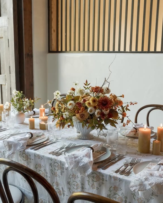 A beautifully styled Thanksgiving table featuring a floral centerpiece with autumn hues, surrounded by elegant dinnerware and warm candlelight, set against a minimalistic backdrop.