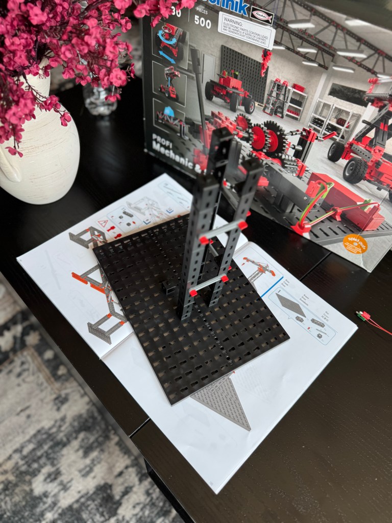 An assortment of Fischer Technik STEM kit pieces arranged on a table, featuring a partially constructed model with a black base and red connectors, alongside the instruction manual and a decorative vase with flowers.