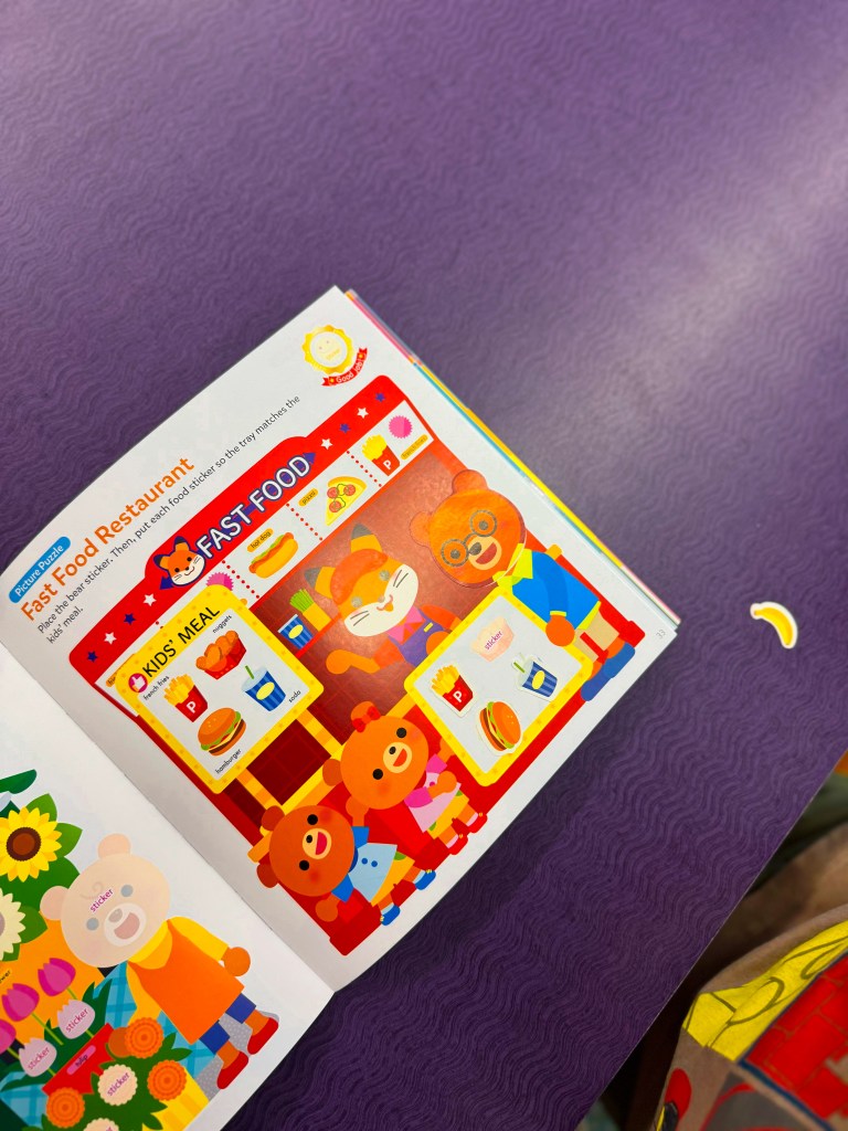 An open children's book page featuring a colorful illustration of a fast food restaurant, including cartoon bears wearing clothing, a menu board, food items like burgers and fries, and a playful atmosphere.