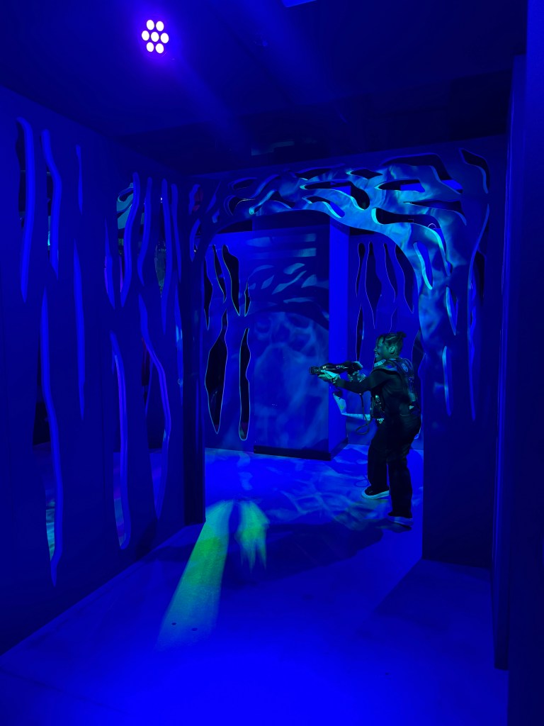 A child dressed in a black vest is aiming a laser gun in a dark, blue-lit laser tag arena with abstract wall designs.