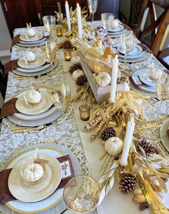 A beautifully styled Thanksgiving table featuring a neutral color palette with mini pumpkins, pinecones, and decorative corn. The setting includes elegant dinnerware, golden accents, and soft candlelight to create a warm and inviting atmosphere.