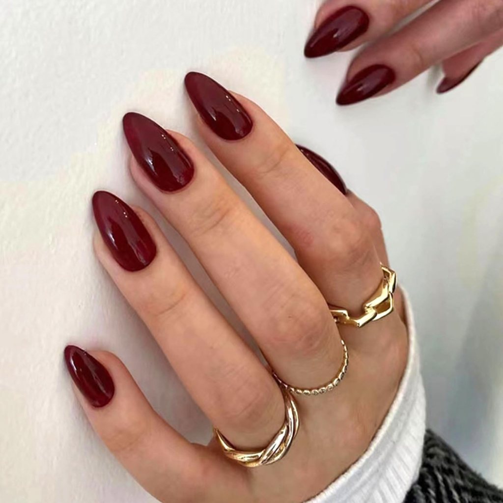 Close-up of hands with long, almond-shaped burgundy nails and gold rings against a neutral background.