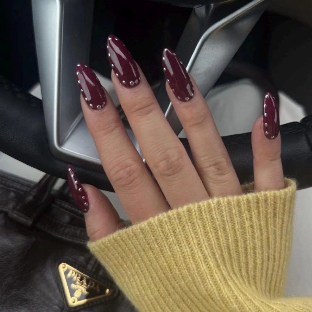 Close-up of a hand with long, burgundy nails decorated with silver studs, resting on a steering wheel, and wearing a yellow sweater.