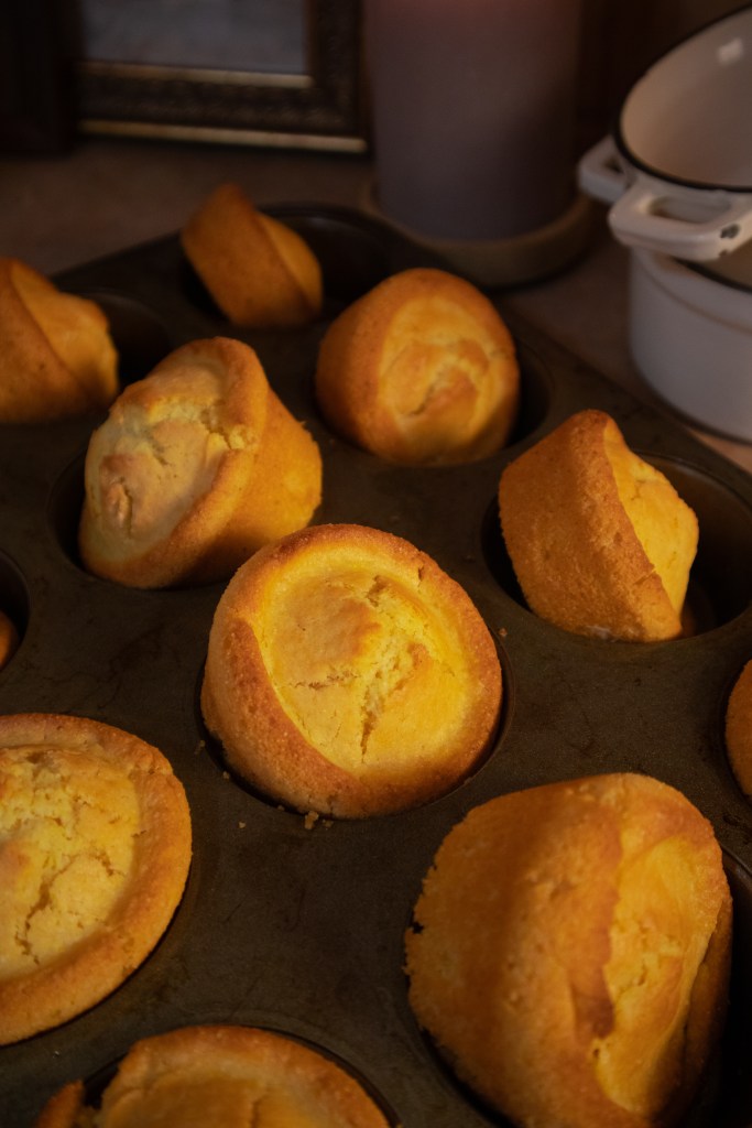 A freshly baked tray of cornbread muffins in a dark metal muffin pan, with some muffins rounded and others slightly collapsed.