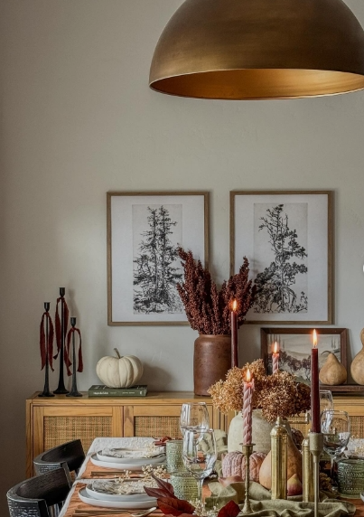 A cozy fall dining table setting featuring soft pastel colors, decorated with pumpkins, candles, and elegant tableware. The warm atmosphere is complemented by dried florals and framed botanical art, creating an inviting ambiance.