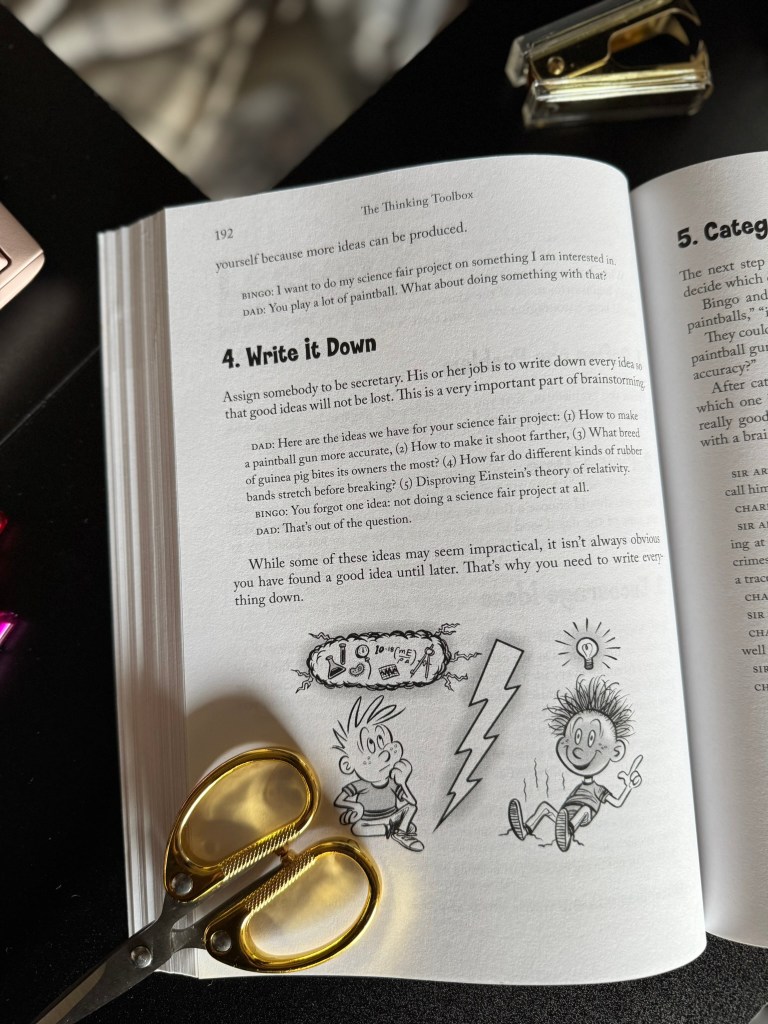 An open page of 'The Thinking Toolbox' book displaying the text '4. Write it Down' with cartoon illustrations of children brainstorming and a pair of scissors in the foreground.