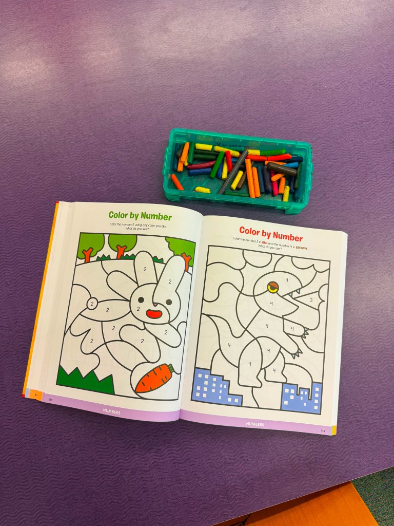 Open color by number books featuring a bunny and a dinosaur, with numbered sections to fill in. Nearby, a green container holds various colorful crayons.