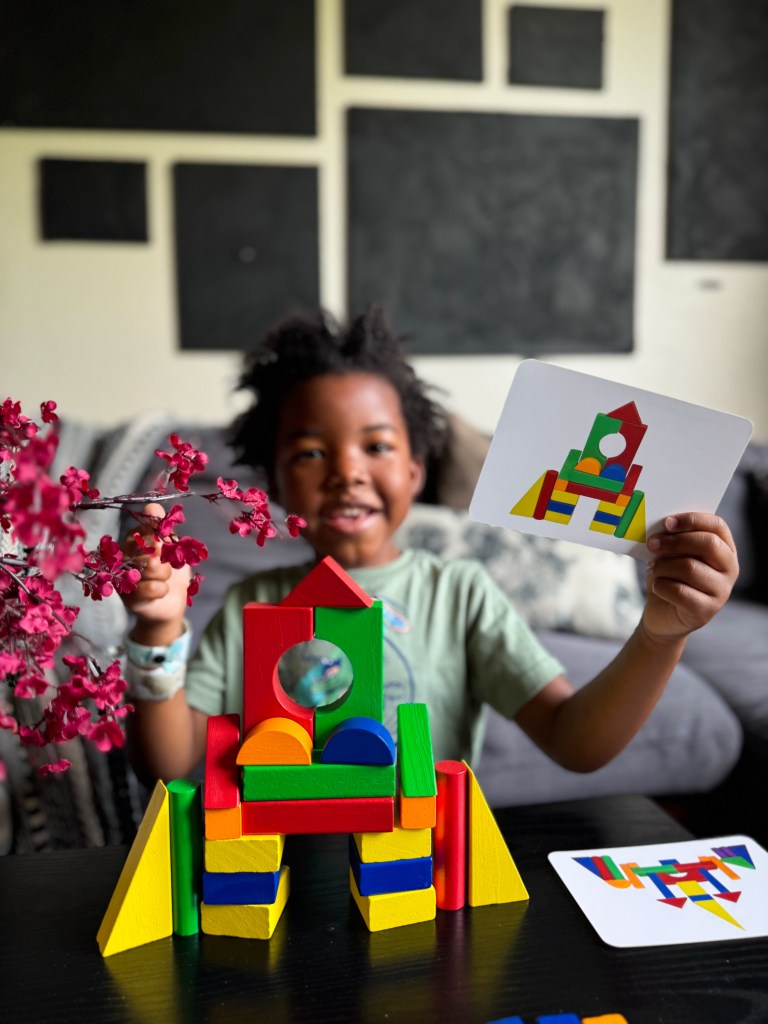A child smiles while holding a card and showing a colorful structure made of building blocks, with blooming flowers in the foreground.