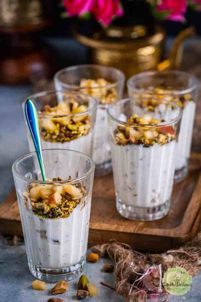 Six clear glasses filled with creamy dessert topped with nuts and chopped apples, placed on a wooden tray.