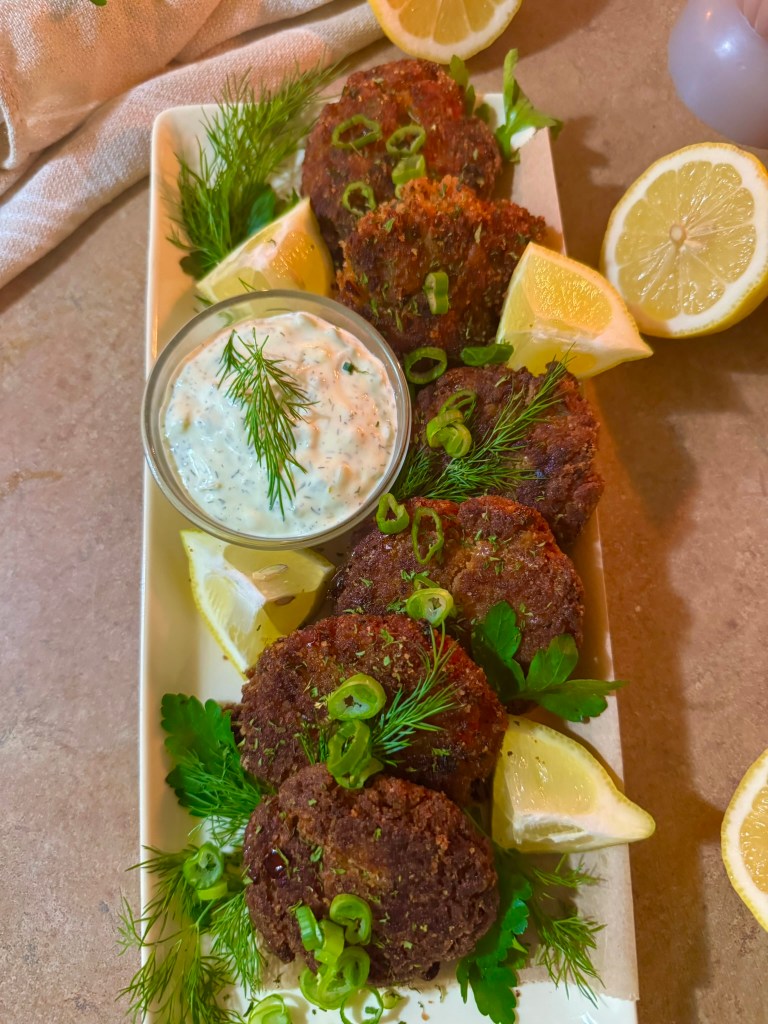 A platter of crispy salmon croquettes garnished with green onions and fresh herbs, served with a lemon wedge and a side of dill dip.