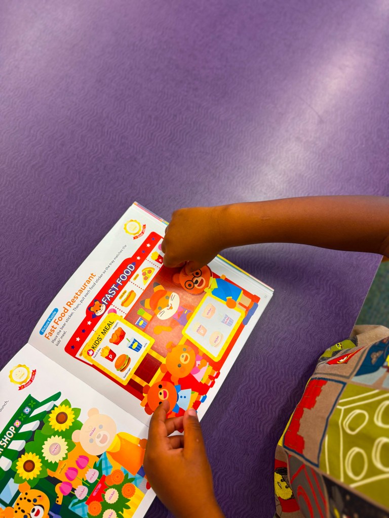 A child's hands interacting with a colorful illustrated page from a storybook about a fast food restaurant, featuring cute animal characters and food items, on a purple table.