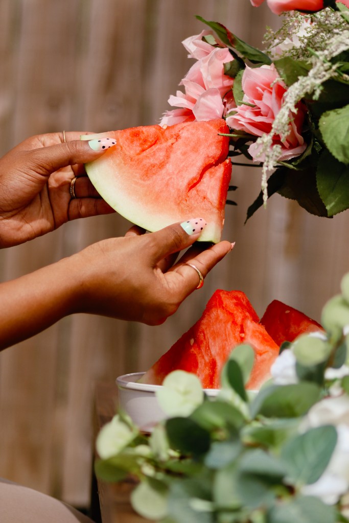 A person holding a slice of watermelon near a bouquet of pink flowers and greenery.