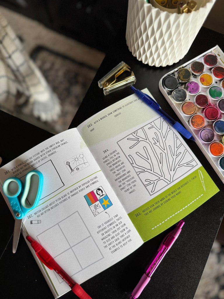 An open art book featuring creative prompts, surrounded by various art supplies including scissors, pens, and a watercolor palette, on a dark tabletop.