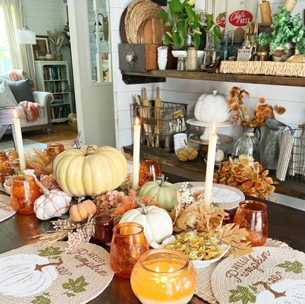 A cozy fall tablescape featuring pumpkins, candles, and autumn leaves arranged on a wooden table. The setup includes soft pastel colors, decorative plates, and a warm ambiance created by candlelight.