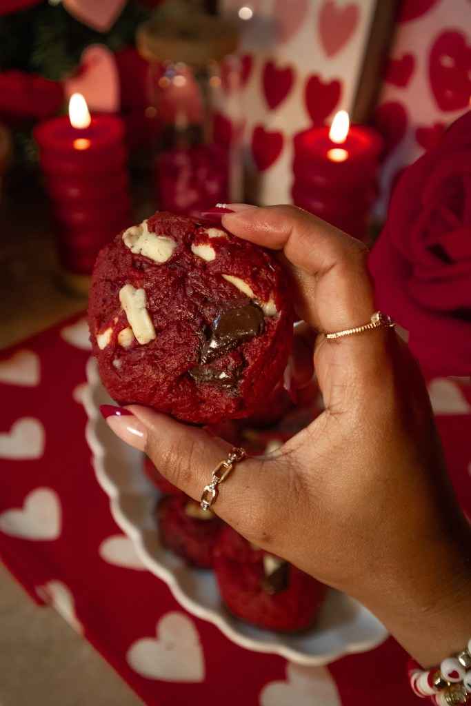 A hand holding a red velvet cookie with chocolate chips, set against a romantic backdrop of candles and heart-themed decorations.