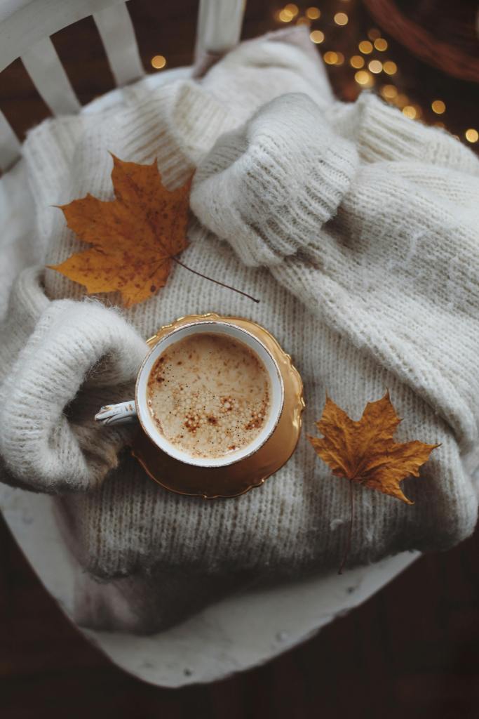 A cozy scene featuring a warm cup of coffee or latte placed on a knitted sweater, accompanied by fall leaves, creating a snug autumn atmosphere.