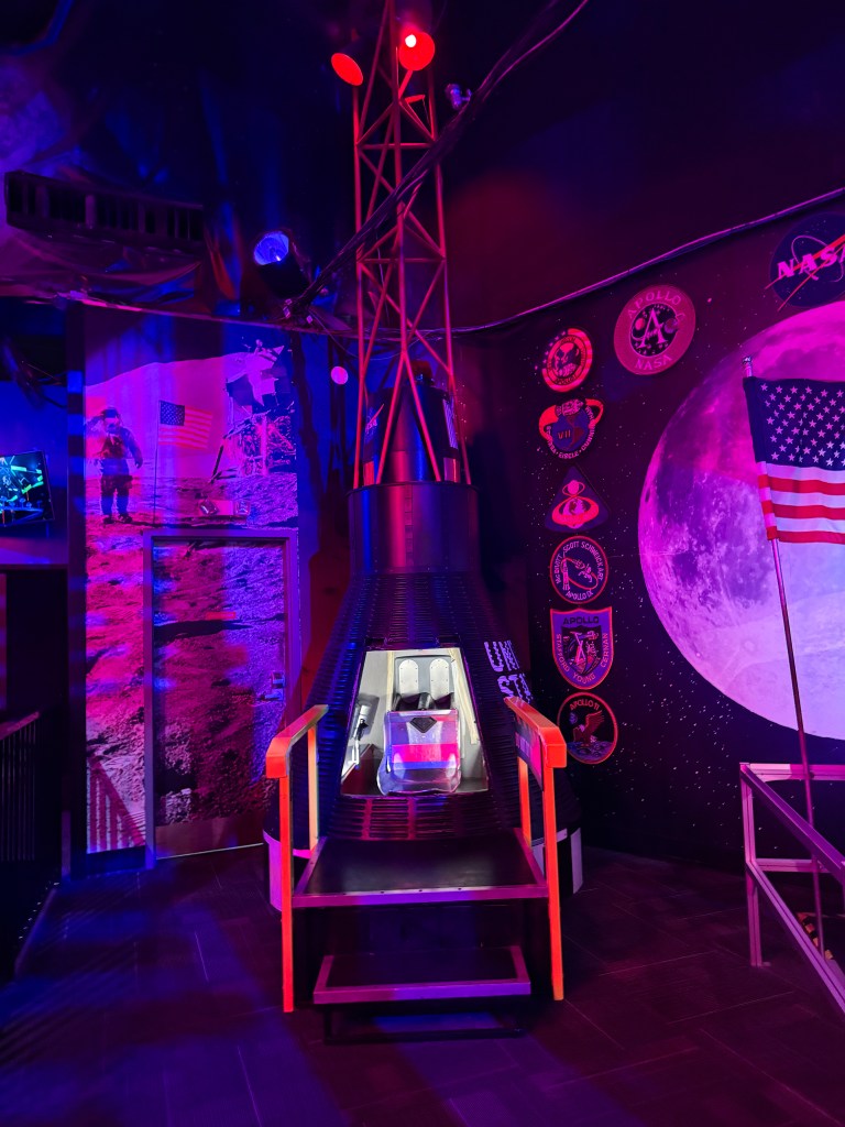 A futuristic spacecraft model with a detailed cockpit, illuminated with colorful lights. It features a metal frame with stairs and is set against a backdrop of space imagery and NASA insignias.
