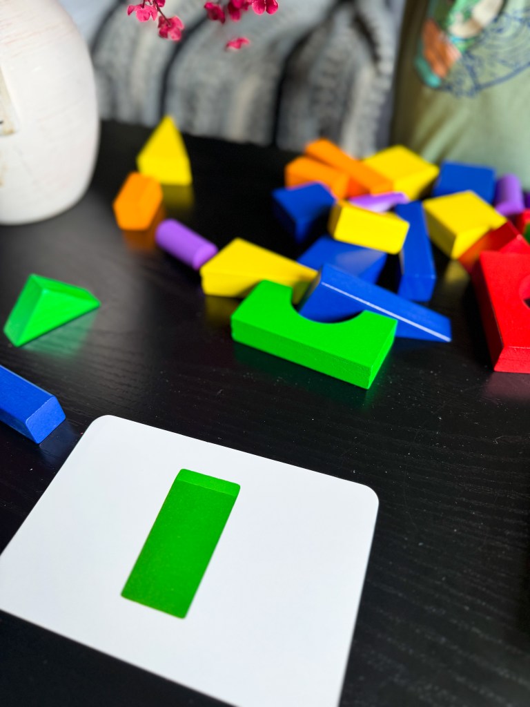 A colorful assortment of geometric blocks scattered on a black table, with a white card displaying a cut-out shape in the center.