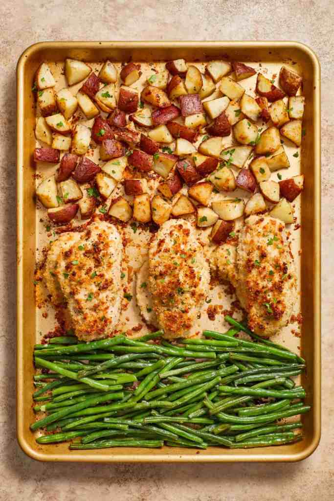 A sheet pan meal featuring crispy baked chicken breasts topped with breadcrumbs, served alongside roasted red potatoes and sautéed green beans.